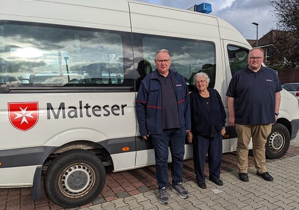 Auf dem Bild ist ein Malteser-Fahrzeug mit zwei Ehrenamtlichen Helfern und einer alten Damen, die als Fahrgast im Mobilen Einkaufswagen mitfährt. 